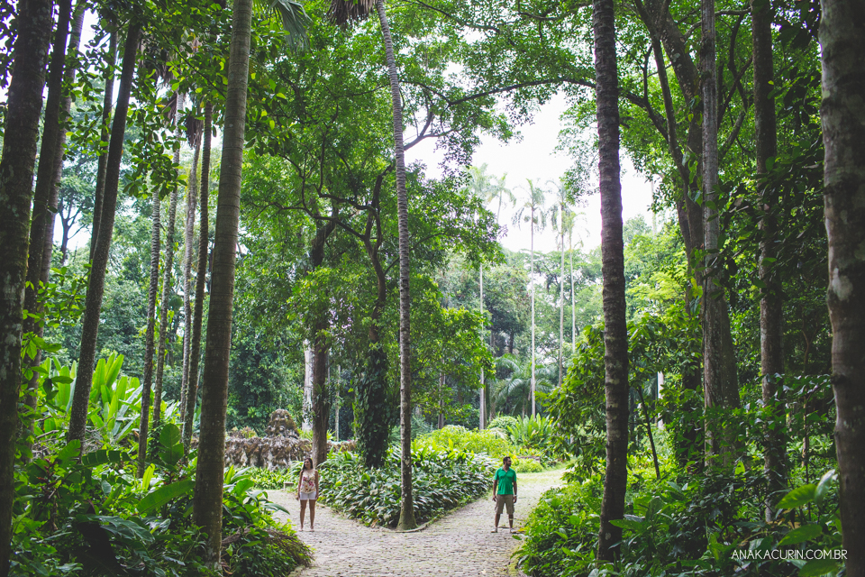 Casal em pé cada um em um caminho de uma bifurcação com longas e majestosas árvores em torno da estrada, emoldurando-os. Fotografia de casal feita por Ana Kacurin no Rio de Janeiro - RJ.