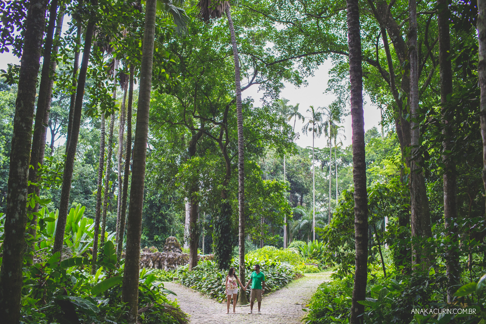 Casal em pé um ao lado do outro, de mãos dadas, em um caminho de uma bifurcação com longas e majestosas árvores em torno da estrada, emoldurando-os. Fotografia de casal feita por Ana Kacurin no Rio de Janeiro - RJ.
