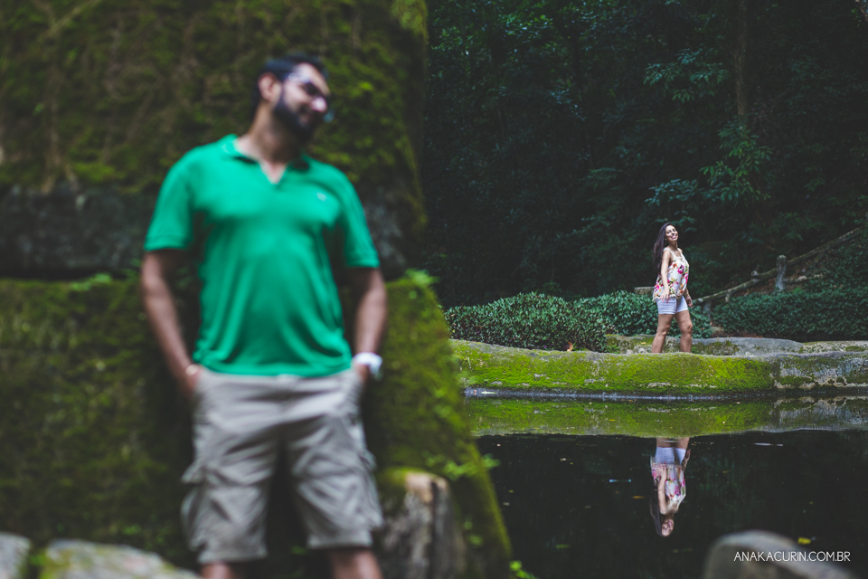Homem recostado em muro de pedra coberto de musgo verde em desfoque, enquanto sua noiva dança ao longe na beira de um lago. Fotografia de casal feita por Ana Kacurin no Rio de Janeiro - RJ.