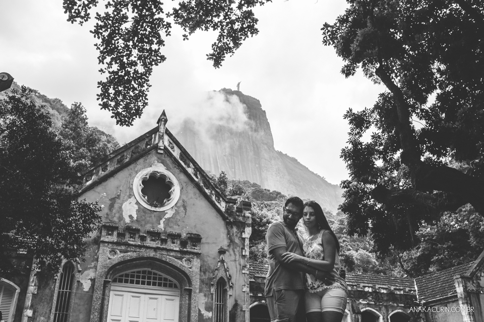 Casal se abraça em frente a uma bela  e majestosa construção antiga. Acima deles, está o Cristo Redentor. Fotografia de casal feita por Ana Kacurin no Rio de Janeiro - RJ.