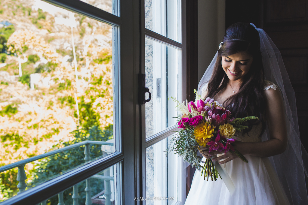 Noiva Larissa olhando seu bouquet, pronta em seu vestido de casamento, próximo à janela