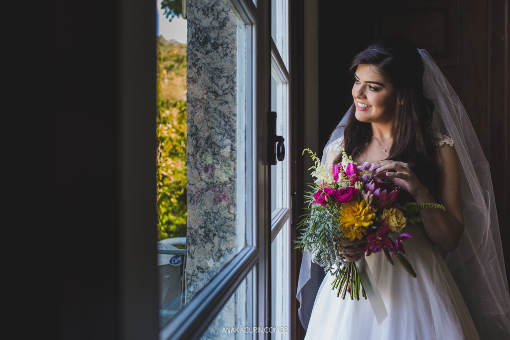 Noiva Larissa olhando seu bouquet, pronta em seu vestido de casamento, próximo à janela