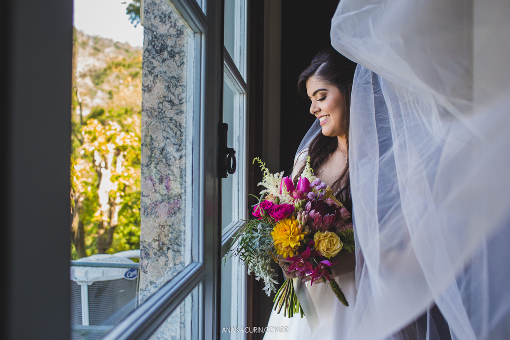 Noiva Larissa olhando seu bouquet, pronta em seu vestido de casamento, próximo à janela