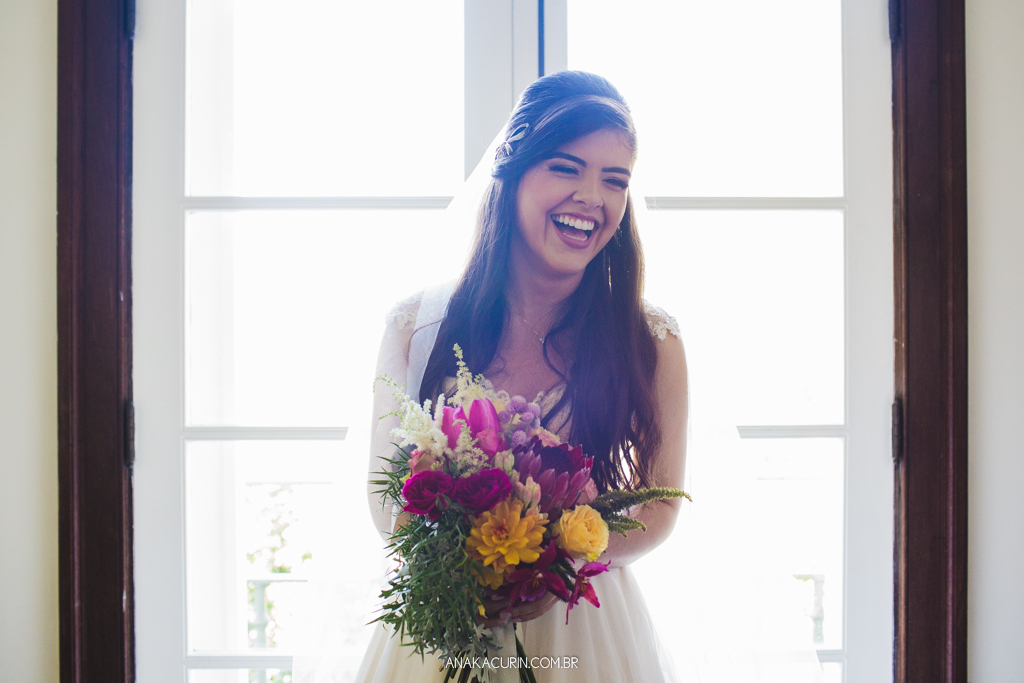 Noiva Larissa olhando seu bouquet, pronta em seu vestido de casamento, próximo à janela, dando uma gostosa gargalhada