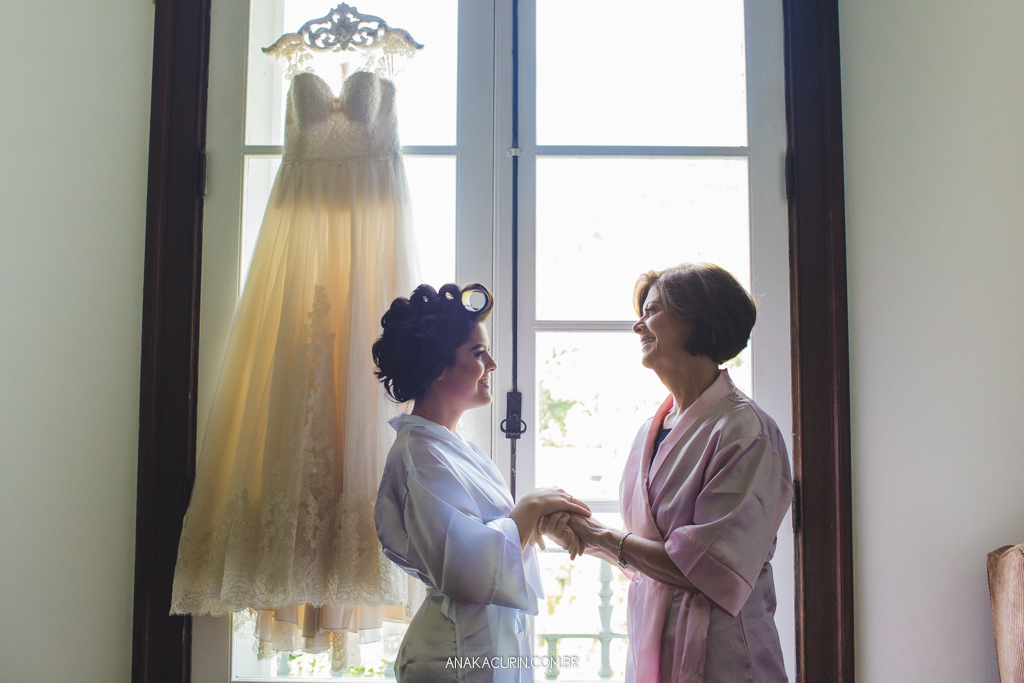 Noiva e sua mãe se abraçam ternamente em frente à janela onde seu vestido está pendurado no contra-luz