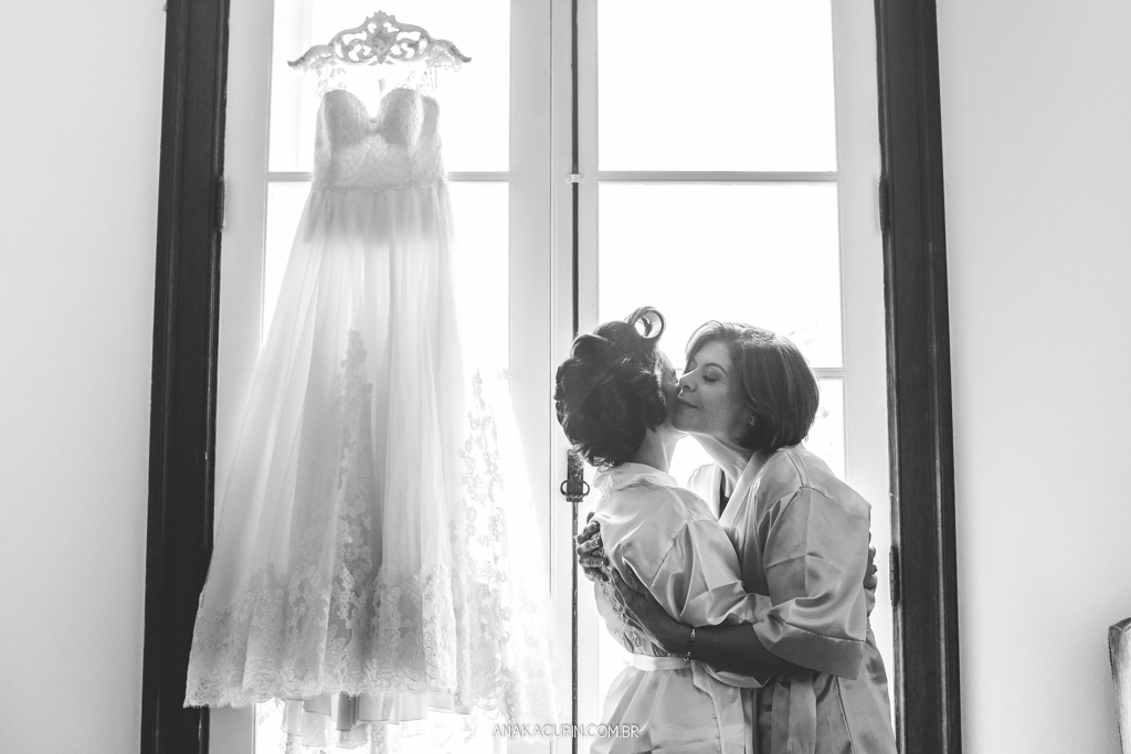 Noiva e sua mãe se abraçam ternamente em frente à janela onde seu vestido está pendurado no contra-luz