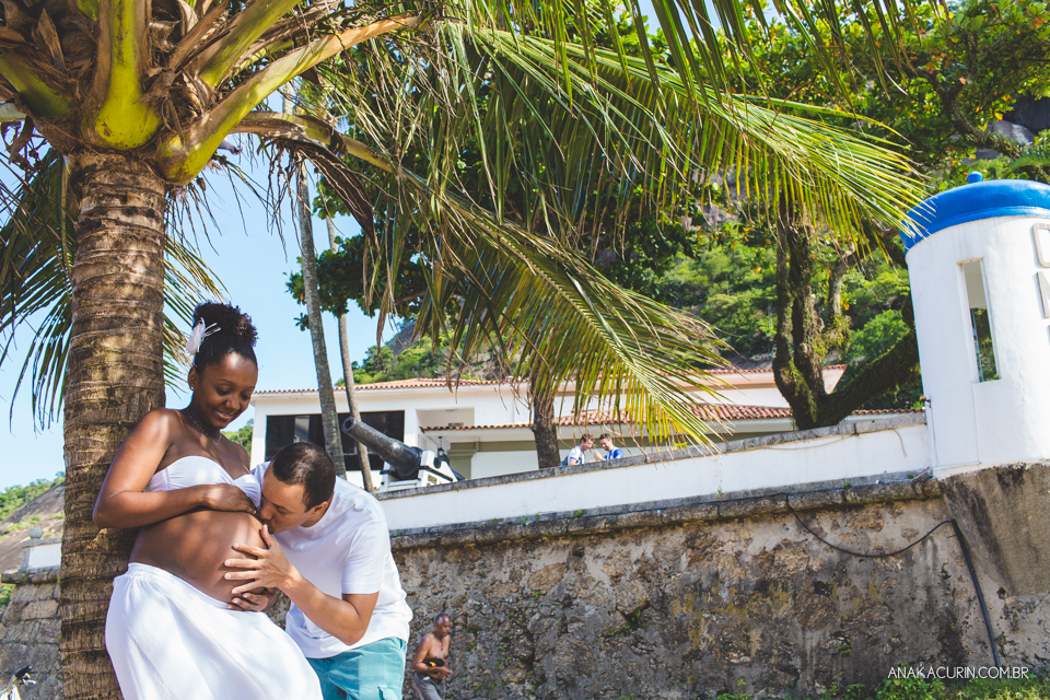 Casal grávido recostado em um coqueiro, iluminado por raios de sol que passa por entre as folhas daa árvores. Ele beija a barriga dela