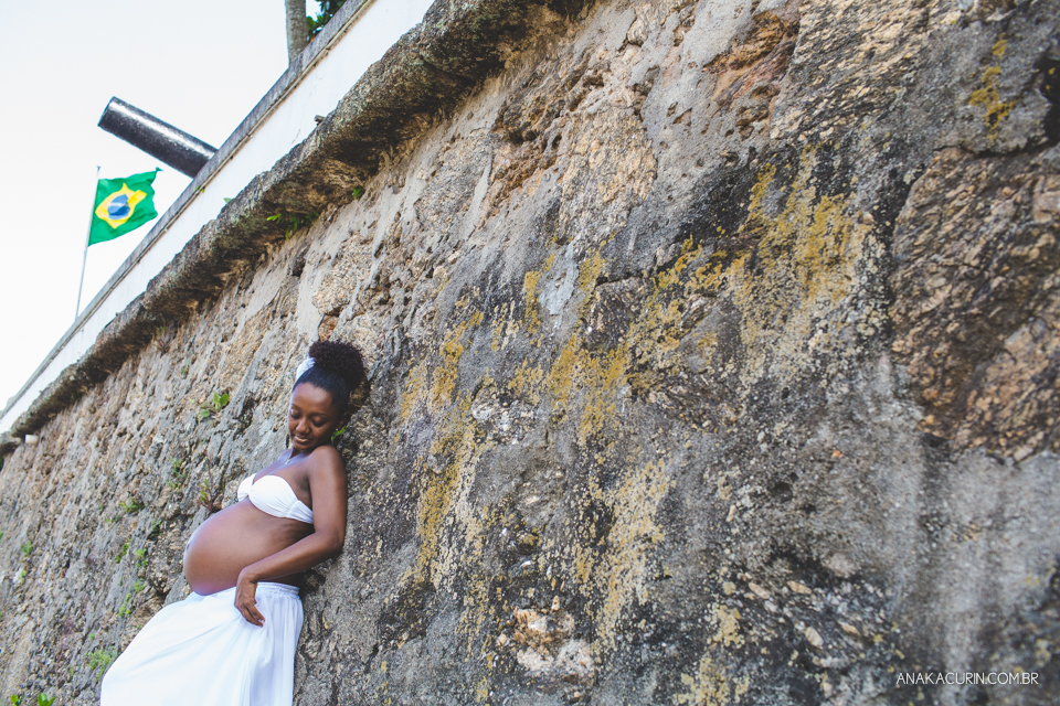 Grávida recostada em parede de pedra, segurando sua saia. Bandeira do Brasil flamulando em mastro ao fundo