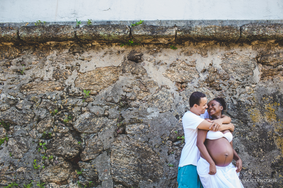Casal grávido abraçado com parede de pedra ao fundo. Ela o olha ternamente