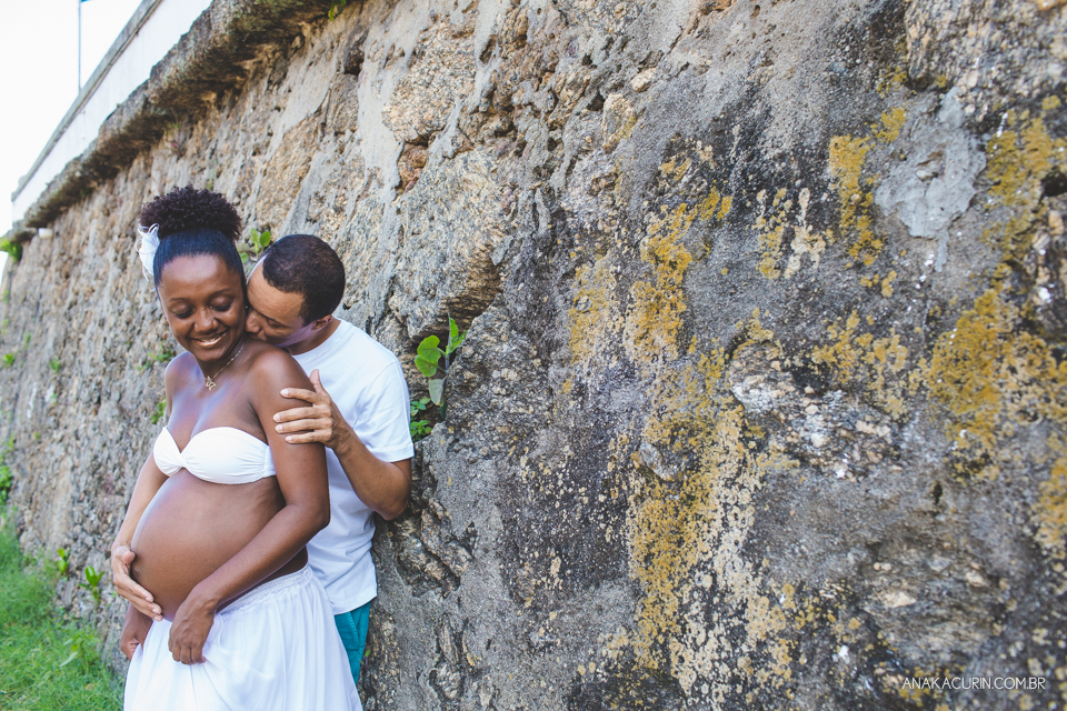 Casal grávido abraçado com parede de pedra ao fundo. Ele beija o pescoço dela com carinho