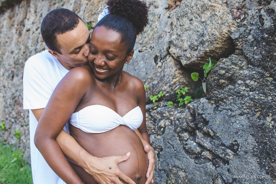 Casal grávido abraçado com parede de pedra ao fundo. Ele beija o rosto dela com carinho