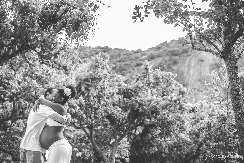 Casal grávido se abraçando com árvores ao fundo