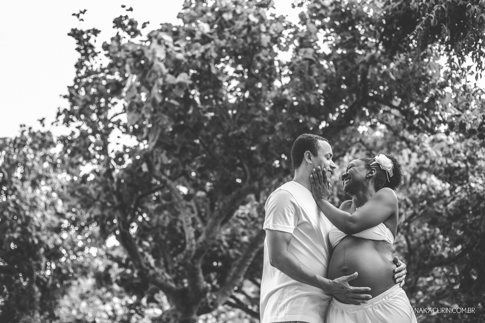 Mulher grávida e risonha toca o rosto do marido ternamente, árvores ao fundo do cenário