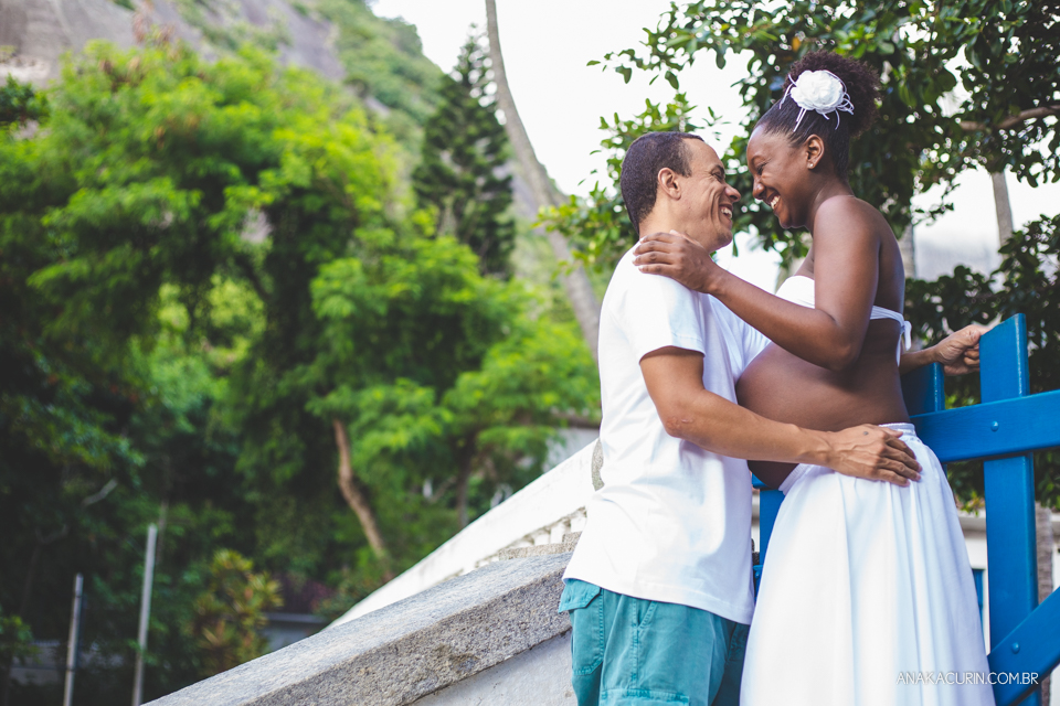 Casal grávido se entreolhando e sorrindo ao topo de uma escadaria com um portão azul ao fundo