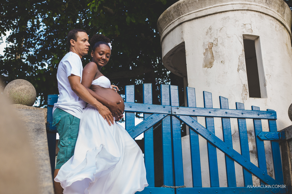 Casal grávido se entreolhando e sorrindo ao topo de uma escadaria com um portão azul ao fundo