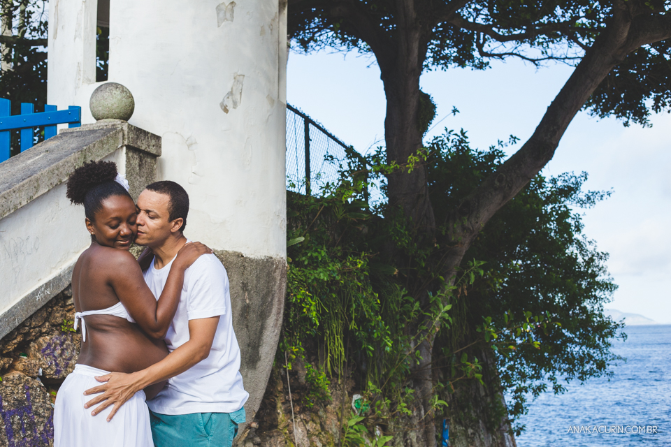 Casal grávido se abraça aos pés da escada com a praia ao fundo, ele a beija no rosto