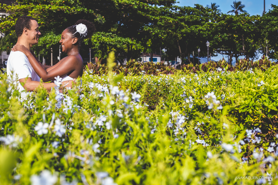 Casal se abraça em meio a uma plantação de flores ensolarada. Céu azul ao fundo