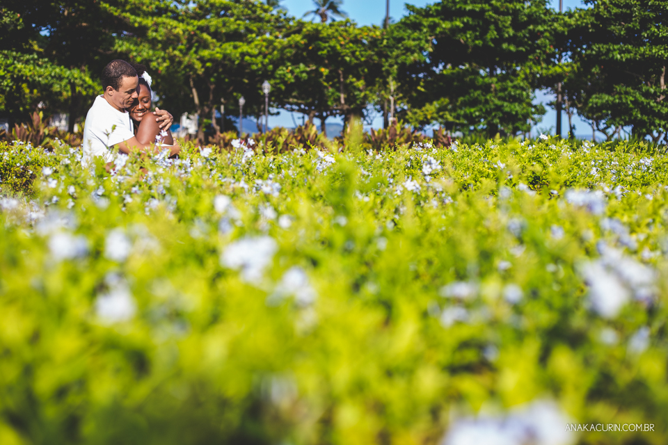 Casal se abraça em meio a uma plantação de flores ensolarada. Céu azul ao fundo