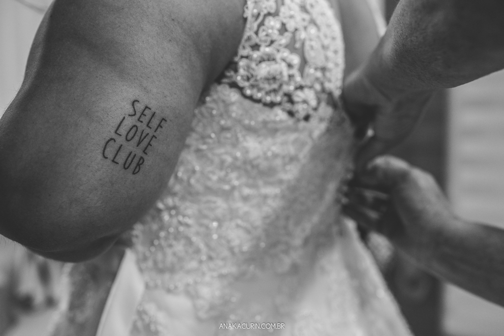 Mãe da noiva ajudando-a a colocar o vestido da estilista Nina Marinho. No braço da noiva, uma tatuagem escrito 