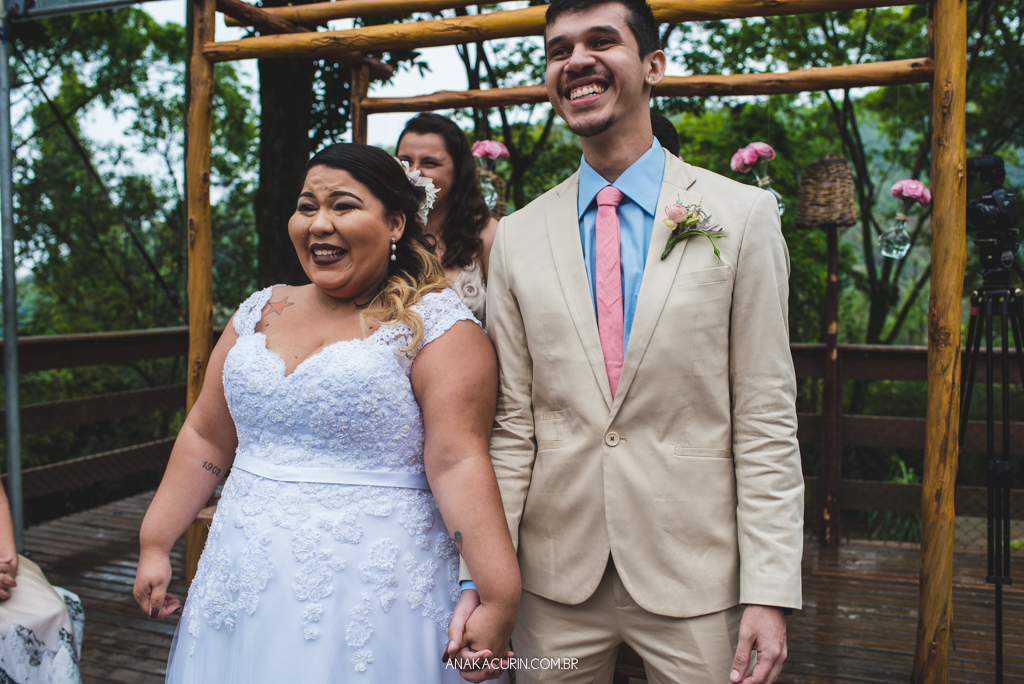 Casal de noivos sorri emocionado ao ver avó da noiva entrar com as alianças em sua cerimônia de casamento