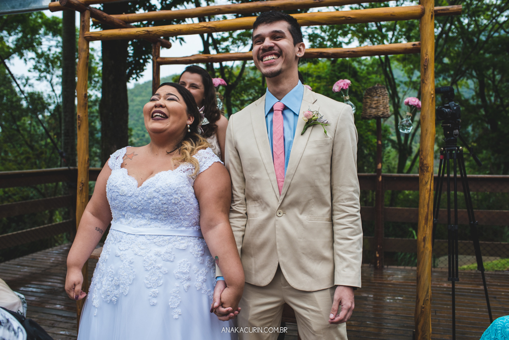 Casal de noivos sorri emocionado ao ver avó da noiva entrar com as alianças em sua cerimônia de casamento