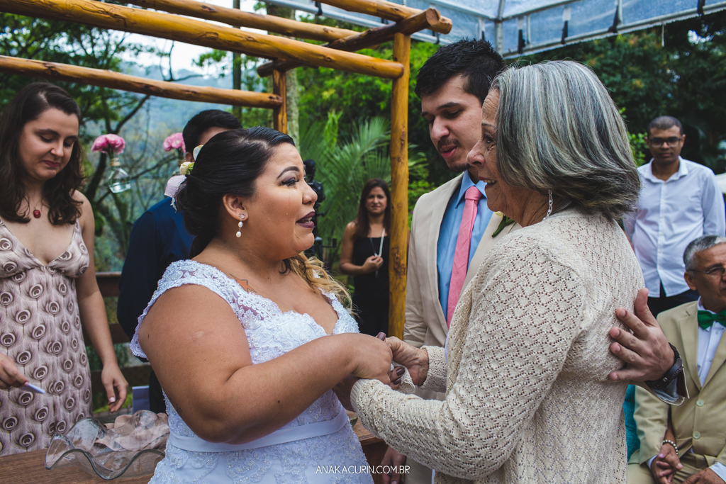 Casal de noivos sorri emocionado ao receber avó da noiva entrar com as alianças em sua cerimônia de casamento