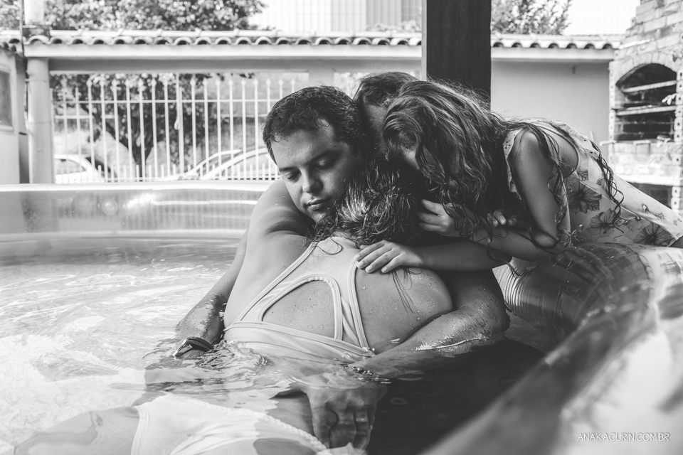 Gestante em trabalho de parto em casa, numa piscina no quintal sendo amorosamente assistida pelas duas filhas pequenas e o marido