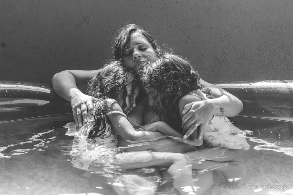 Gestante em trabalho de parto em casa, numa piscina no quintal sendo amorosamente assistida pelas duas filhas pequenas, que a abraçam