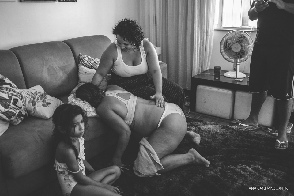 Gestante em trabalho de parto em casa, ajoelhada com a cabeça apoiada no colo de sua doula. Suas duas filhas brincam na sala em volta dela.