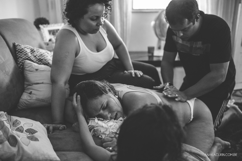 Gestante em trabalho de parto em casa, ajoelhada com a cabeça apoiada no colo de sua doula. Suas duas filhas brincam na sala em volta dela.