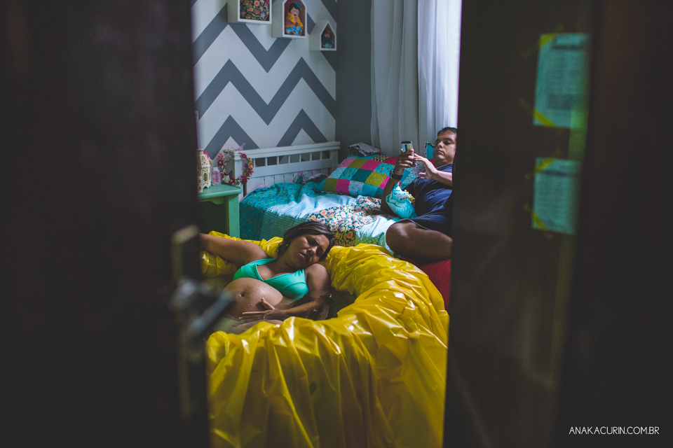 Gestante em trabalho de aprto em casa, mergulhada numa piscina amarela. Seu marido mexe ao celular ao fundo, sentado numa cama de criança.