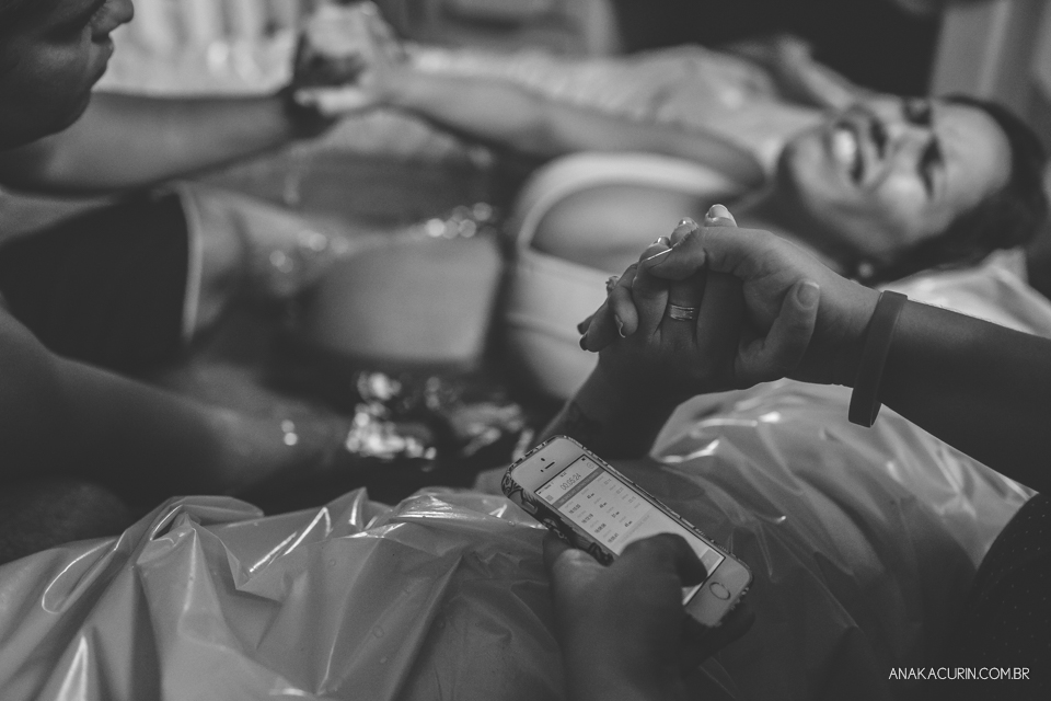 Gestante em trabalho de aprto em casa, mergulhada numa piscina amarela, sendo assistida pelo marido, que a massageia. Foco está nas mãos da gestante, que segura ternamente a mão da doula, que não aparece na foto. Ao lado de sua