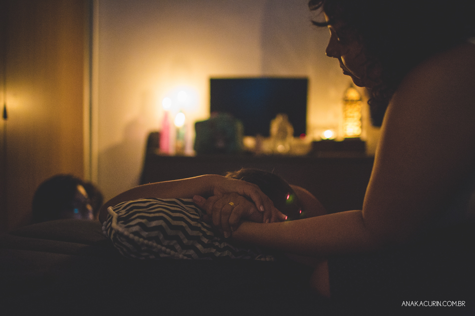 Gestante em trabalho de aprto em casa, num quarto escuro iluminado pela luz de velas, sendo assistida pela parteira, doula e o marido