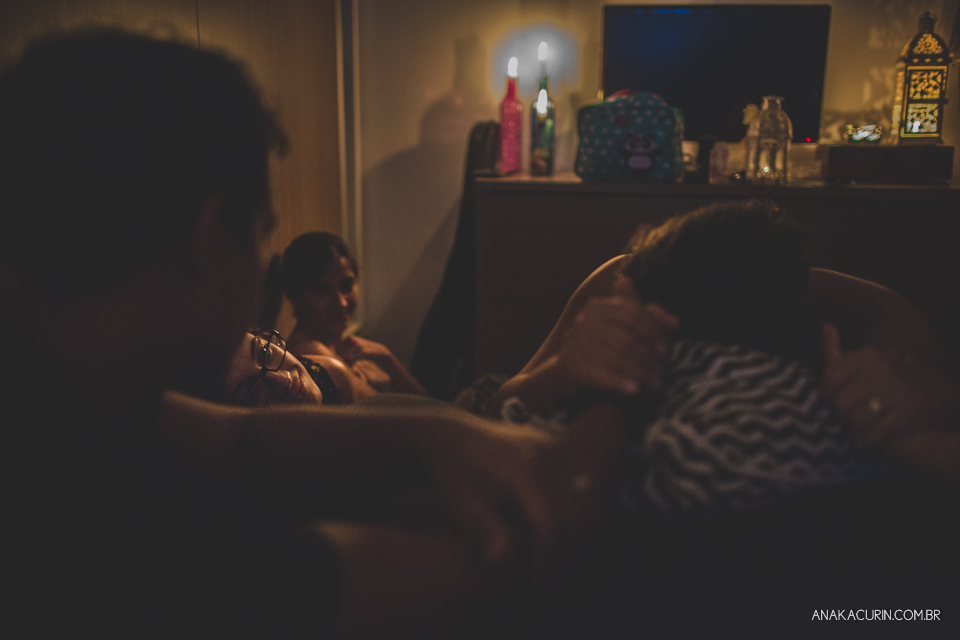 Gestante em trabalho de aprto em casa, num quarto escuro iluminado pela luz de velas, sendo assistida pela parteira, doula e o marido