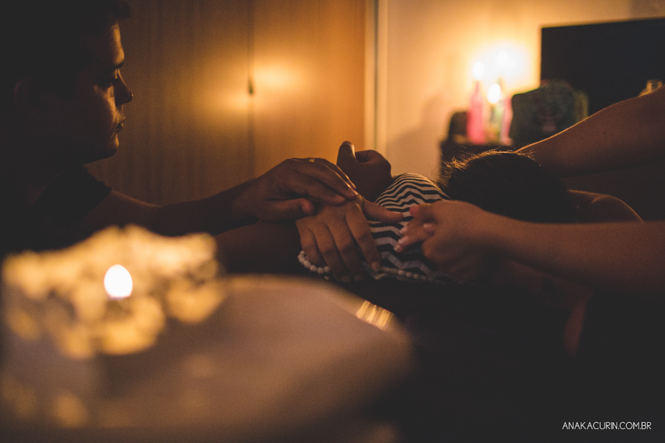 Gestante em trabalho de aprto em casa, num quarto escuro iluminado pela luz de velas, sendo assistida pela parteira, doula e o marido