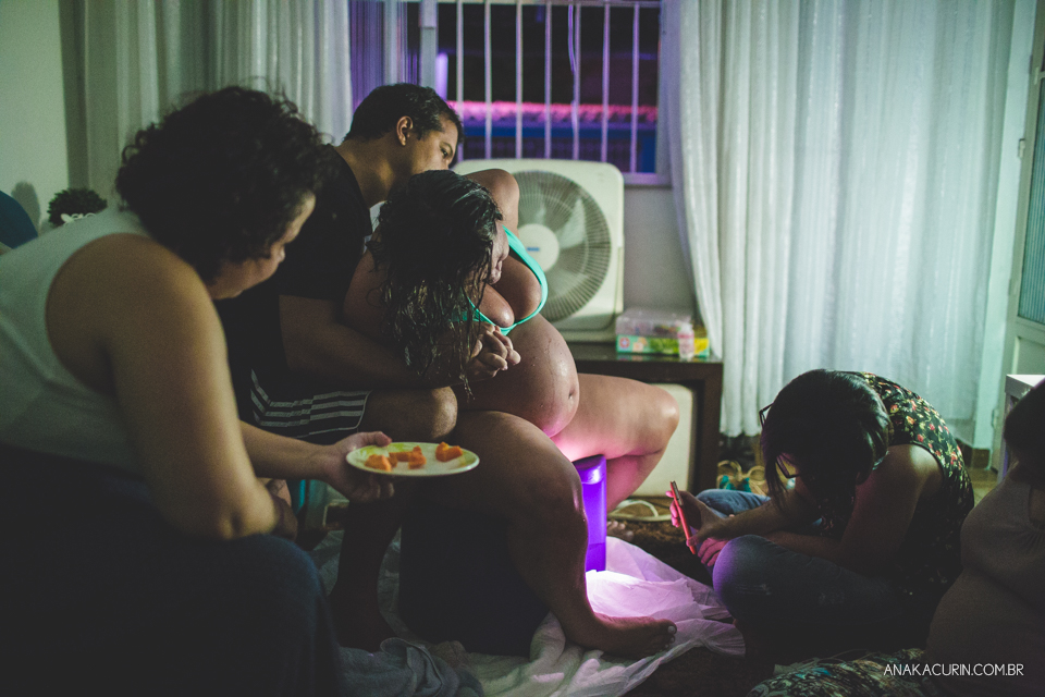 Gestante em trabalho de parto em casa sentada na banqueta de parto, sendo amorosamente sustentada pelo marido. Doula lhe oferecendo frutas cortadinhas.