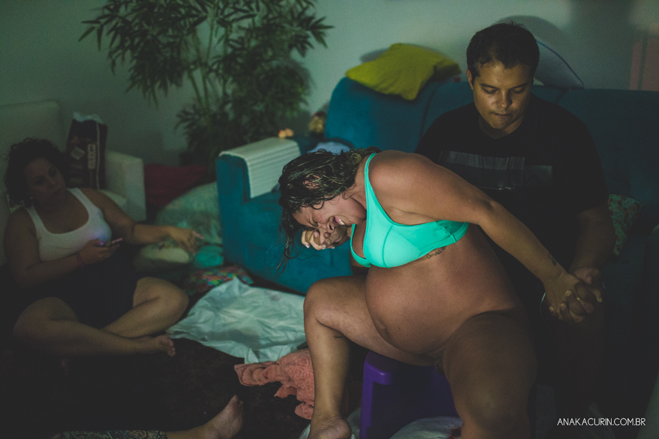 Gestante em trabalho de parto em casa sentada na banqueta de parto com expressão de dor, sendo amorosamente sustentada pelo marido.  parteira sentada à sua frente observando-a.