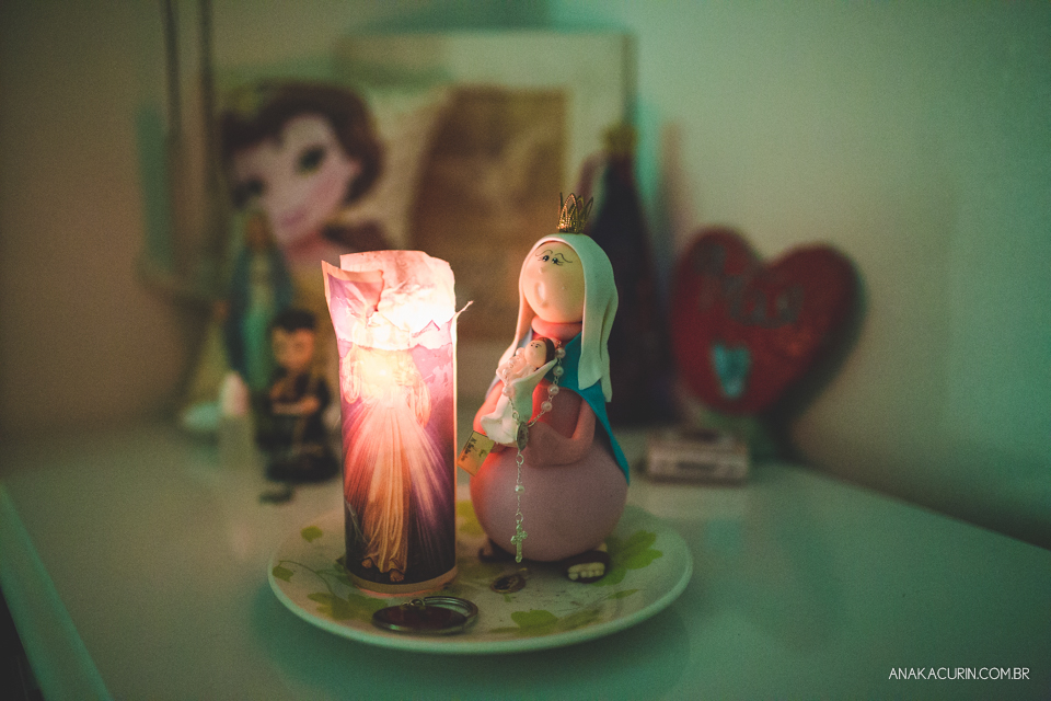 Imagem de nossa senhora do parto feita em biscuit ao lado de vela acesa. Ao fundo, coisas de bebê.