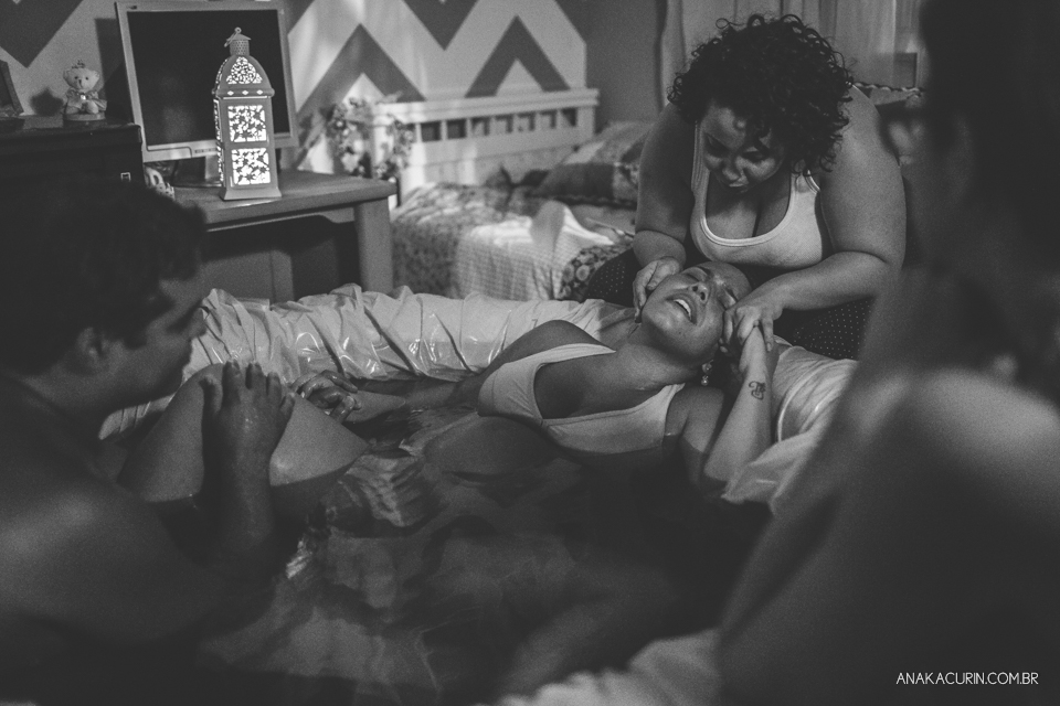 Gestante parindo em piscina em sua casa, sendo amparada por sua doula e marido, e assistida por sua parteira