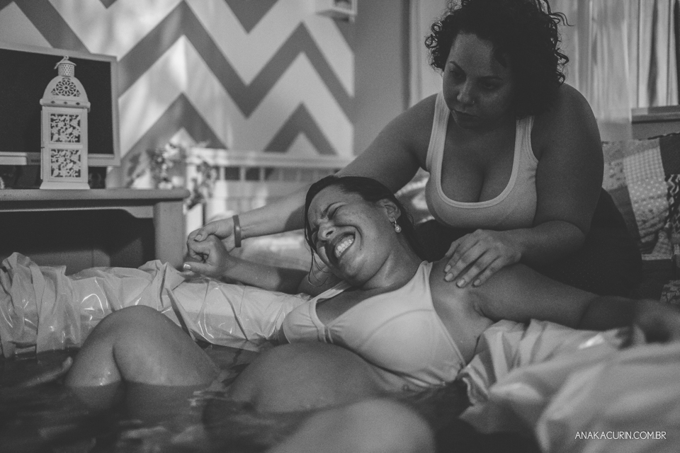 Gestante parindo em piscina em sua casa, sendo amparada por sua doula e marido, e assistida por sua parteira