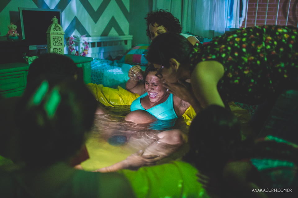 Gestante parindo em piscina em sua casa, sendo amparada por sua doula e marido, e assistida por sua parteira. Poide-se ver a cabeça do bebê já do lado de fora.