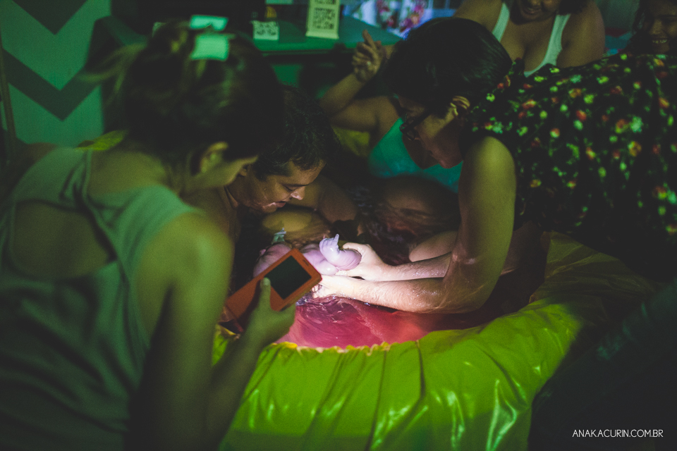 Parteira e pai recebem o bebê na piscina de casa enquanto gestante ao fundo é amparada pela doula