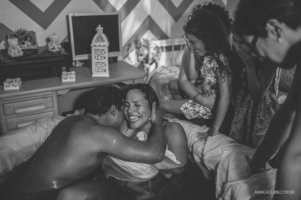 Pai beija rosto de gestante com bebê recém parido nos braços, dentro da piscina de casa. As duas filhas do casal choram ao fundo, emocionadas.