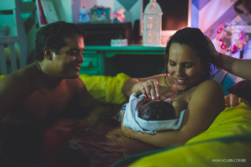 mulher e marido olham ternamente para bebê recém parido no colo, dentro da piscina, em casa.