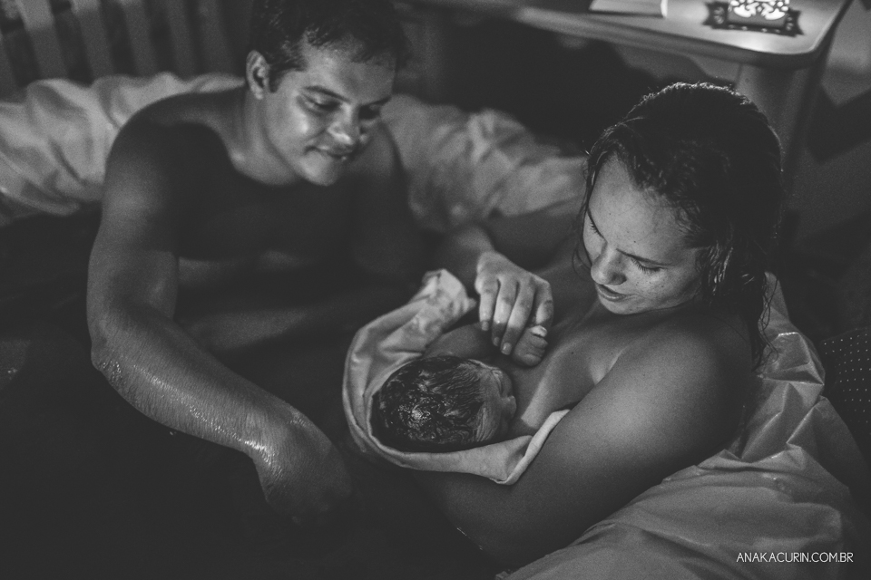 mulher e marido olham ternamente para bebê recém parido no colo, dentro da piscina, em casa.