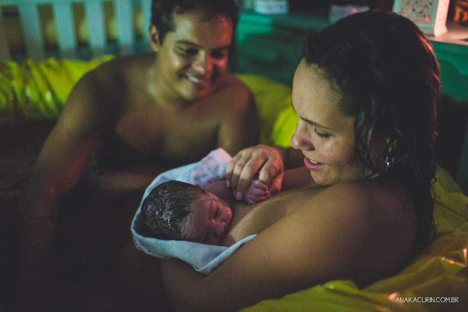 mulher e marido olham ternamente para bebê recém parido no colo, dentro da piscina, em casa.