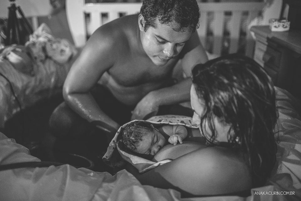 mulher e marido olham ternamente para bebê recém parido no colo, dentro da piscina, em casa.