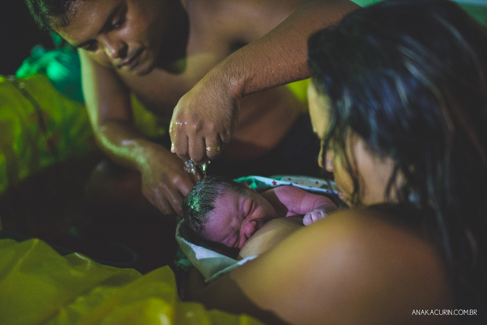 mulher e marido olham ternamente para bebê recém parido no colo, dentro da piscina, em casa.