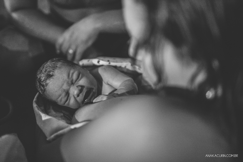 mulher e marido olham ternamente para bebê recém parido no colo, dentro da piscina, em casa.