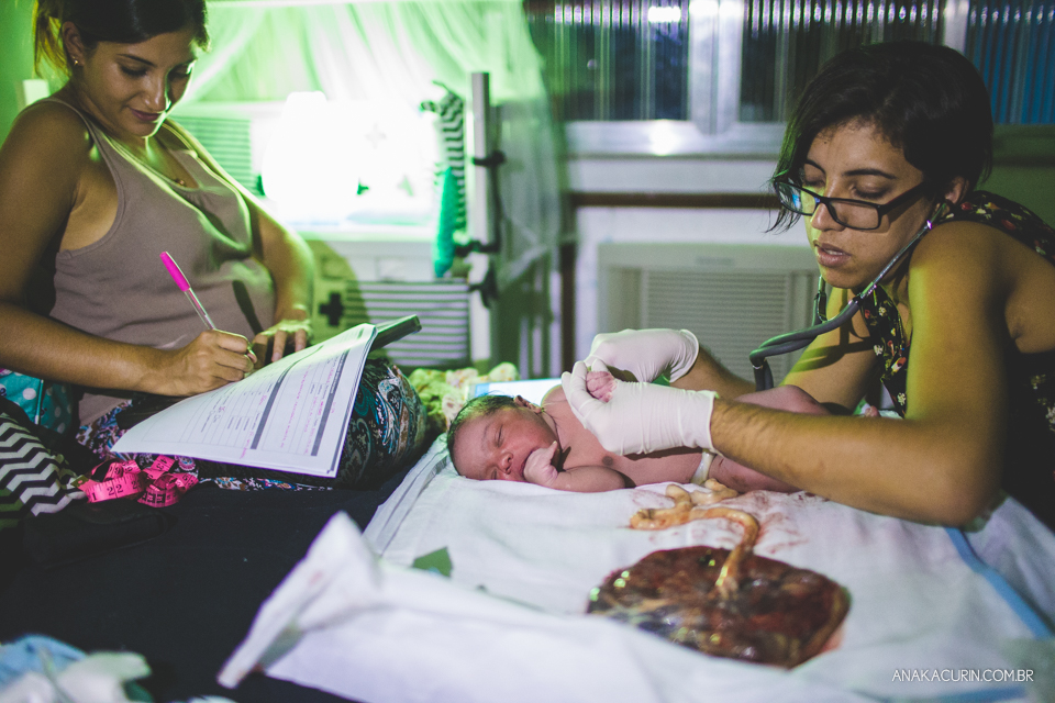 bebê recém nascido ainda conectado À pacenta através do cordão umbilical, sendo examinado pela parteira
